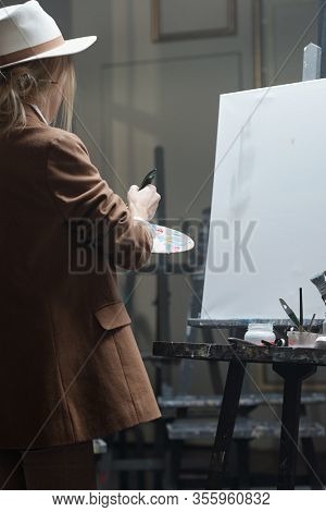 Young woman in hat and jacket mixing colors on palette while standing in front of easel with blank paper and going to paint