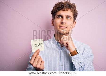 Young blond romantic man with curly hair holdiing paper with love message for valentines day serious face thinking about question, very confused idea