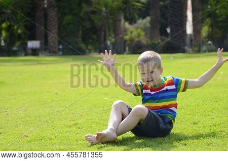 Israel Rishon Lezion 22 May 2022 Positive Child Sits On The Green Grass In The Park. Boy In A Multi-