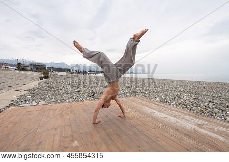 Shirtless Caucasian Man Doing Backflip On Pebble Beach.