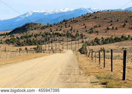 Rural Countryside Dirt Road Besides Rustic Fences At Ranchlands Taken On Arid Badlands With Snow Cap