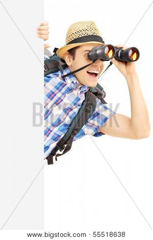 Smiling male tourist loooking through binocular behind a blank panel isolated on white background