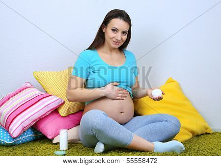 Young regnant woman sitting on floor and applying cream on her belly on wall background