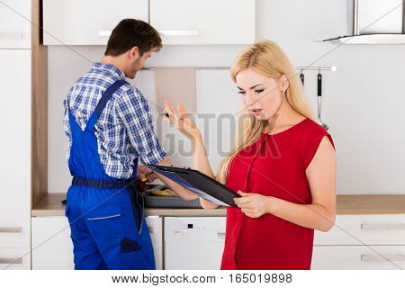 Young Frustrated Woman Looking At Expensive Bill On Clipboard With Plumber Standing In Kitchen
