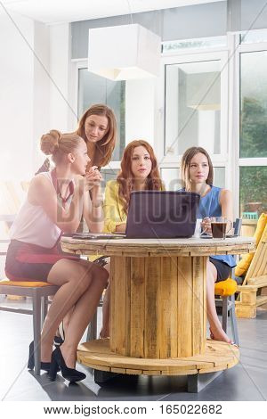 Group of four pretty businesswomen working togeather with new startup project using laptop computer in modern loft. People and teamwork concept - happy creative team in office. Casually dressed female colleagues talking in a meeting room.