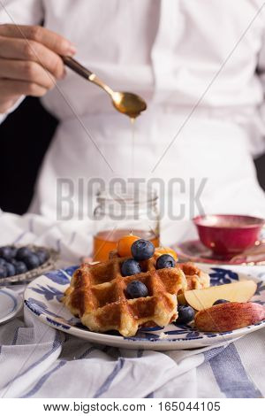 Blueberry apple waffle with honey set on a dish with a chef's hand holding spoon for a jar of honey ready to serve.