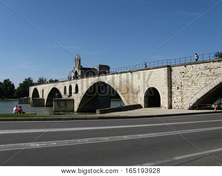 Medieval stone Benezet bridge admire by sitting couple in french historical city of AVIGNON with desolate road in FRANCE and clear blue sky in warm sunny summer day, PROVENCE, EUROPE, JUNE