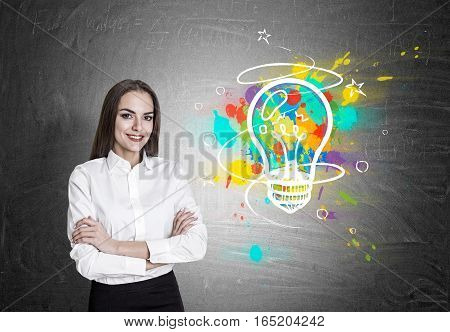 Portrait of a cheerful businesswoman standing near a blackboard with her arms crossed. There is a colorful light bulb drawing on it
