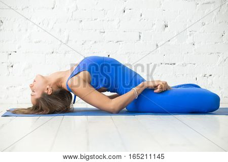 Young attractive woman practicing yoga, sitting in Fish exercise, variation of Matsyasana with lotus legs pose, working out, wearing sportswear, blue suit, indoor full length, white studio background