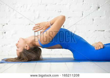 Young attractive woman practicing yoga, stretching in Fish exercise, variation of Matsyasana with lotus legs pose, Namaste, working out, wearing sportswear, blue suit, indoor, white studio background