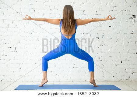 Young attractive woman practicing yoga, standing in Goddess exercise, Sumo Squat pose, working out, wearing sportswear, blue suit, indoor full length, white loft studio background, rear view