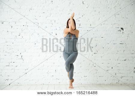 Young attractive woman practicing yoga, standing in Eagle exercise, Garudasana pose, working out, wearing sportswear, grey suit, indoor full length, white studio background