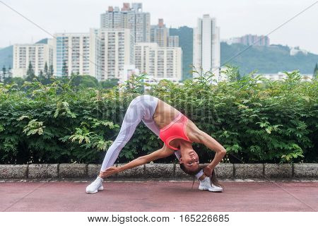 Sporty young woman stretching doing yoga triangle pose standing in wide-legged position bending to side. Fitness girl exercising in the city park in summer.