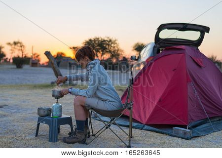 Woman cooking with gas stove in camping site at dusk. Gas burner pot and smoke from boiling water tent in the background. Adventures in african national parks.