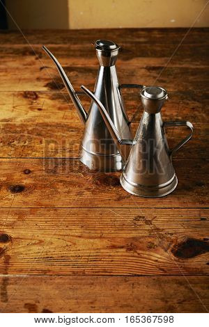 Two similar shaped steel olive oil cruets in a rustic vintage beige toned kitchen on aged wooden table presented close together