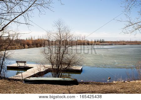 Two canoes laying at a partially frozen rivers edge by floating wood dock in spring landscape