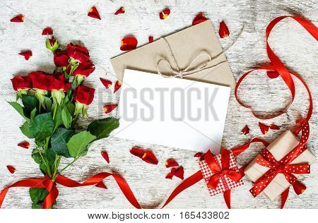 blank white greeting card with red roses bouquet envelope gift boxes and heart shaped red ribbon on white rustic wooden background. top view. valentines day background
