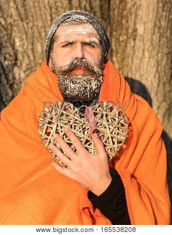 Frozen man with squinty eyes bearded hipster with beard and moustache covered with white frost wrapped in orange blanket with wicker heart on winter day outdoors on natural background