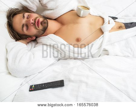 young handsome bearded man in white terry bathrobe sleeping on bed in hotel bedroom with remote control and coffee cup has bare chest