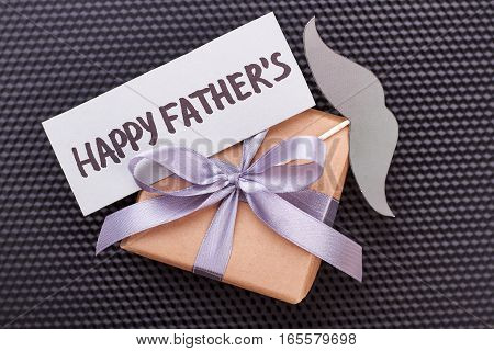 Card and mustache on gift. Present box on black surface. Stylish congratulation for dad.