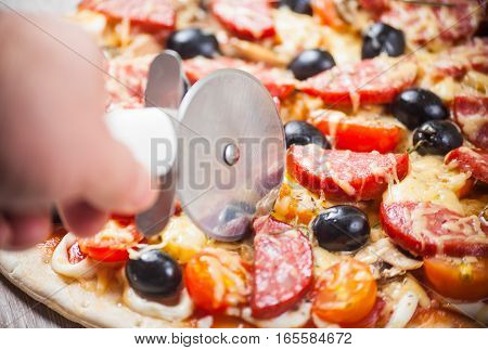 Cook pizza cutter cuts pizza with cheese olives and salami mushrooms and tomatoes. On a wooden stand.