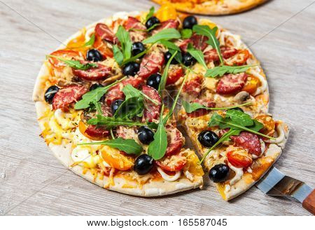 Cook takes a shovel one piece of pizza with cheese olives and salami mushrooms and tomatoes arugula. On a wooden stand.