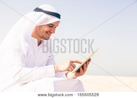 young arabian man sitting at the beach, typing on his tablet computer