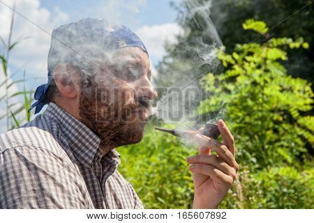Bearded man enjoying his pipe and releasing smoke outside on a summer day