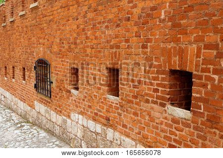 A strong red brick wall surrounding an ancient castle or antique fortress as fortification. The bricks and blocks have different color tones and shades of red. Square holes and a steel grid opening lead into the inner of the medieval structure.
