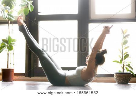 Young attractive woman practicing yoga, stretching in Paripurna Navasana pose, working out, wearing sportswear, grey pants, bra, indoor full length, home interior background, near the floor window