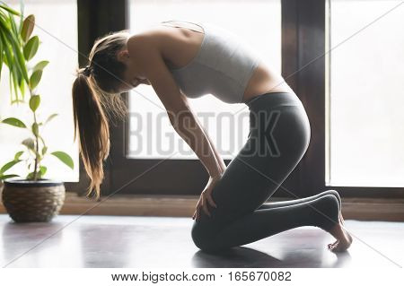 Young attractive woman practicing yoga, sitting in Upward Abdominal Lock exercise, Uddiyana Bandha pose, working out, wearing sportswear, grey pants, bra, indoor full length, home interior background