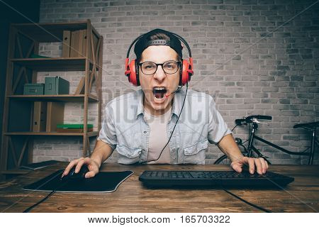Young man playing game at home and streaming playthrough or walkthrough video. Screaming boy in headphones in front of computer trying to fight with his enemies.
