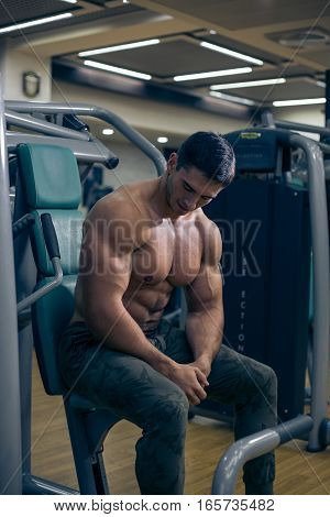 Young bodybuilder looking at the muscles after a workout. Handsome guy sitting on the simulator. Bodybuilder tired after a workout. The man sitting in the gym. A guy with a naked torso. Sexy guy.