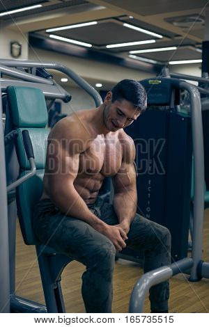 Young bodybuilder looking at the muscles after a workout. Handsome guy sitting on the simulator. Bodybuilder tired after a workout. The man sitting in the gym. A guy with a naked torso. Sexy guy.