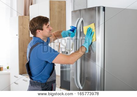Young Man Cleaning Stainless Refrigerator With Rag And Bottle Spray In Kitchen