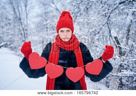 Love and valentines day concept. Discontent sullen woman holding garland of four red paper hearts shape - blank copy space for letters or text, looking at camera over winter landscape