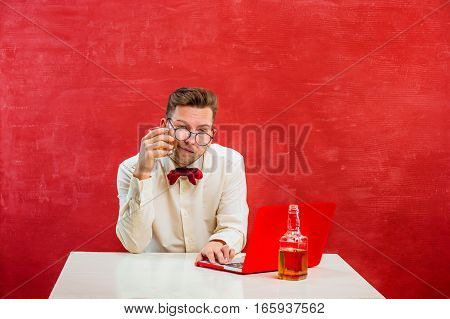 The young funny man with cognac sitting with laptop at St. Valentine's Day on red studio background. Concept - unhappy love