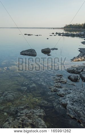 Dead calm morning image of shallow and clear lake huron water and limestone rocks along theshoreline. A small tree-covered peninsula is on the horizon. Overall feeling is a calm peaceful tranquil serene and wild background.