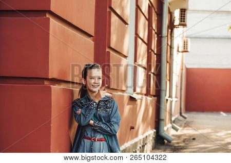 Cheerful Little Girl In Jeans Clothes Stands Near An Old Restored Beautiful Building.