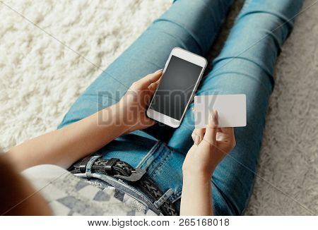 Young Woman Holding Credit Card And Using Phone Sitting On Floor. Payment.  Online Shopping Concept.