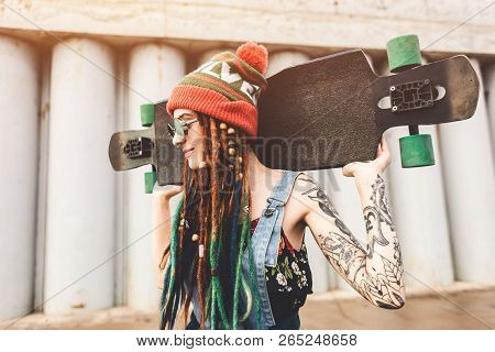 Young Girl In Sun Glasses And A Cap Stands With A Longboard Against The Background Of Concrete Struc