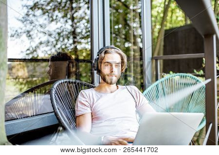 Poortrait Of A Handsome Man Working With Laptop And Headphones On The Balcony Of The House In The Be
