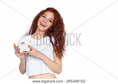 Beautiful Young Woman With Curly Ginger Hair Laughing And Looking At Camera While Holding Piggy Bank