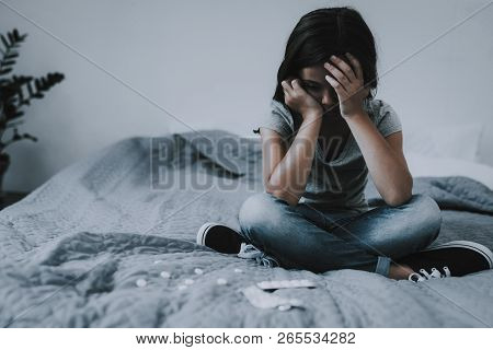 Little Girl Sits On Bed With Scattered Pills. Child On Gray Badcover With Crosssed Legs Near Opened 