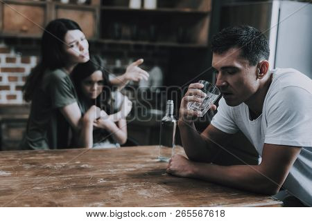 Wife And Kid Look At Father Getting Drunk At Home. Man Sits On Chair In Modern Kitchen Hold Glass Of
