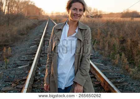 Young Attractive Lady In White Shirt And Beige Raincoat Walking Along The Railroad Tracks Around Blu