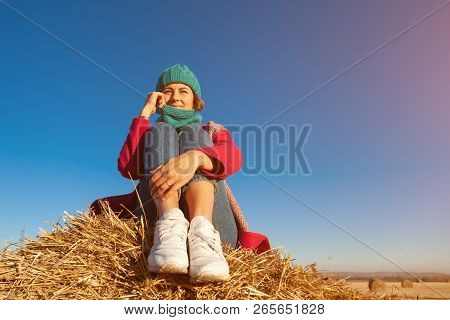 Young Attractive Lady In Blue Knitting Hat And Pink Coat Sitting On Haystack. Smiling Beautiful Hips