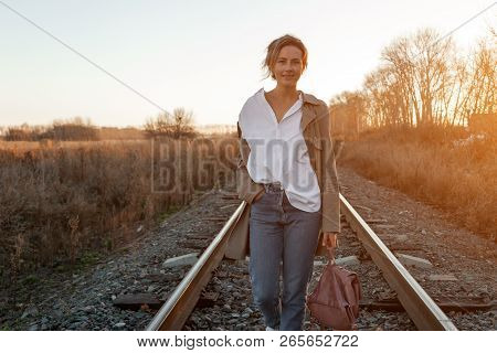 A Young Woman In A White Shirt, Beige Raincoat And Jeans  Enjoys Nature, Walking Along The Railroad 