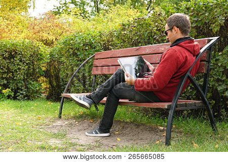 A Young Man In Casual Clothes In A Warm Jacket Working Outdoors In The Autumn Park, Read A Book On U