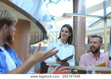 Group Of Young People In Casual Wear Sitting At The Office Desk And Discussing Something While Looki